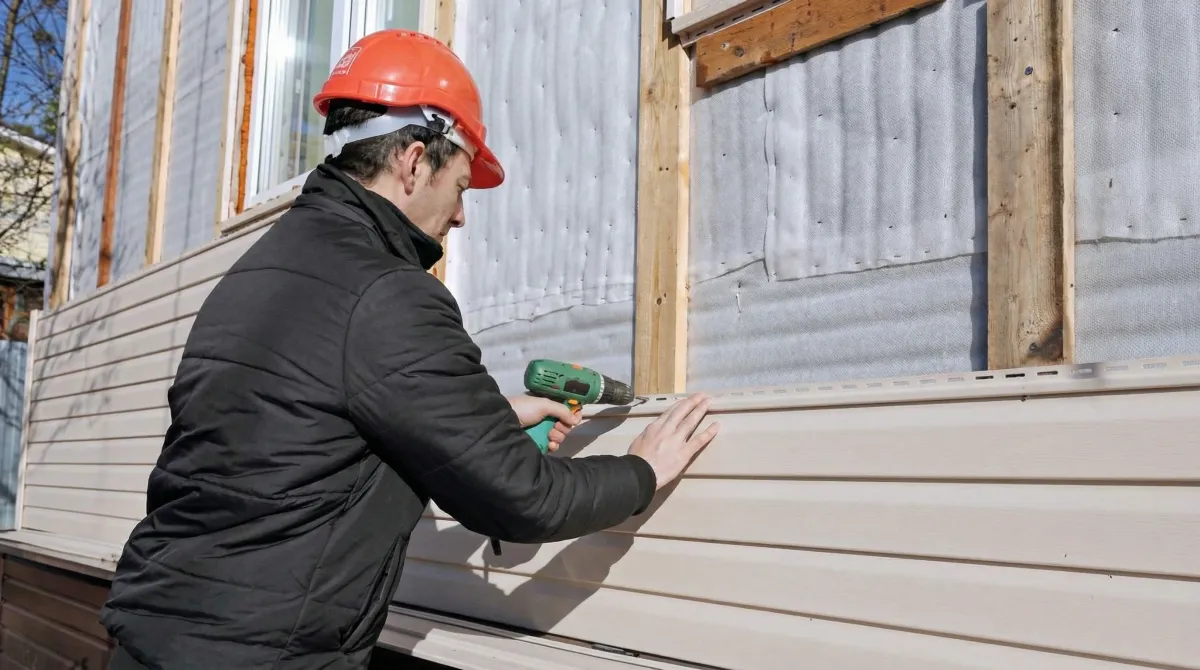 Siding replacement professional using a power drill to install new tan vinyl siding on a home exterior in Providence, RI